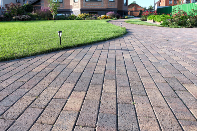 A curved, paved walkway made of rectangular bricks runs through a well-maintained garden with green grass, flowers, and solar path lights near a modern house—expertly crafted by a top masonry contractor in Long Island, NY.
