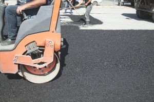 A close-up of a road roller compacting fresh asphalt as workers spread asphalt in the background, showcasing the expertise of a masonry contractor Long Island, NY paving a new road surface under bright daylight.