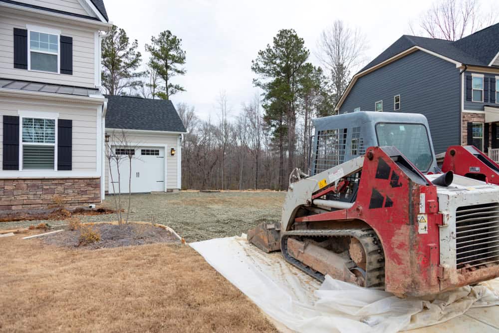 A red and white skid steer loader sits on a plastic-covered driveway in front of a suburban NY house, suggesting work by a masonry contractor Long Island, with another home and trees visible on an overcast day.