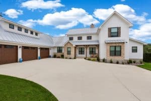 Modern farmhouse in NY with white siding, brick accents by a skilled masonry contractor Long Island, metal roof, three-car garage, and a wide driveway under a blue sky with scattered clouds.