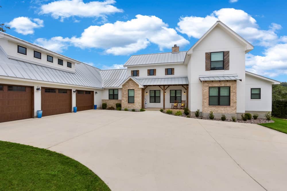 Modern farmhouse in NY with white siding, brick accents by a skilled masonry contractor Long Island, metal roof, three-car garage, and a wide driveway under a blue sky with scattered clouds.