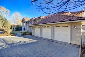 Stunning large luxury house in NY with a red tile roof, elegant columns, expertly crafted by a top masonry contractor Long Island. Features three white garage doors, manicured landscaping, and a paved driveway under a clear blue sky.