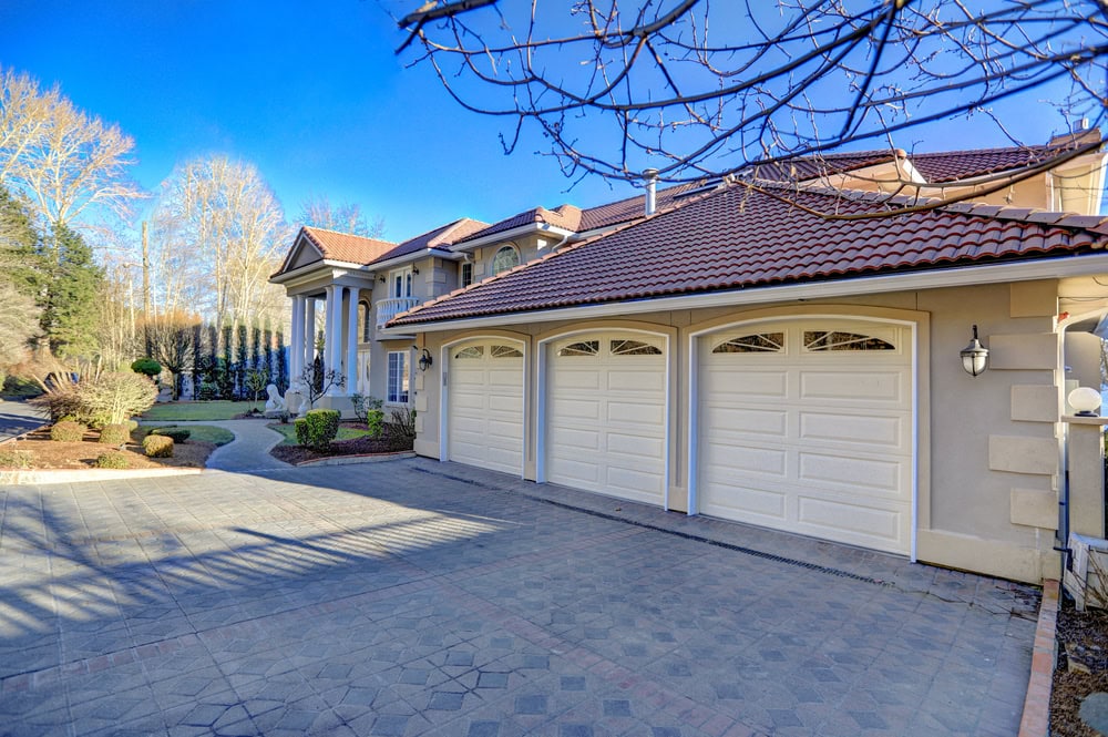 Stunning large luxury house in NY with a red tile roof, elegant columns, expertly crafted by a top masonry contractor Long Island. Features three white garage doors, manicured landscaping, and a paved driveway under a clear blue sky.