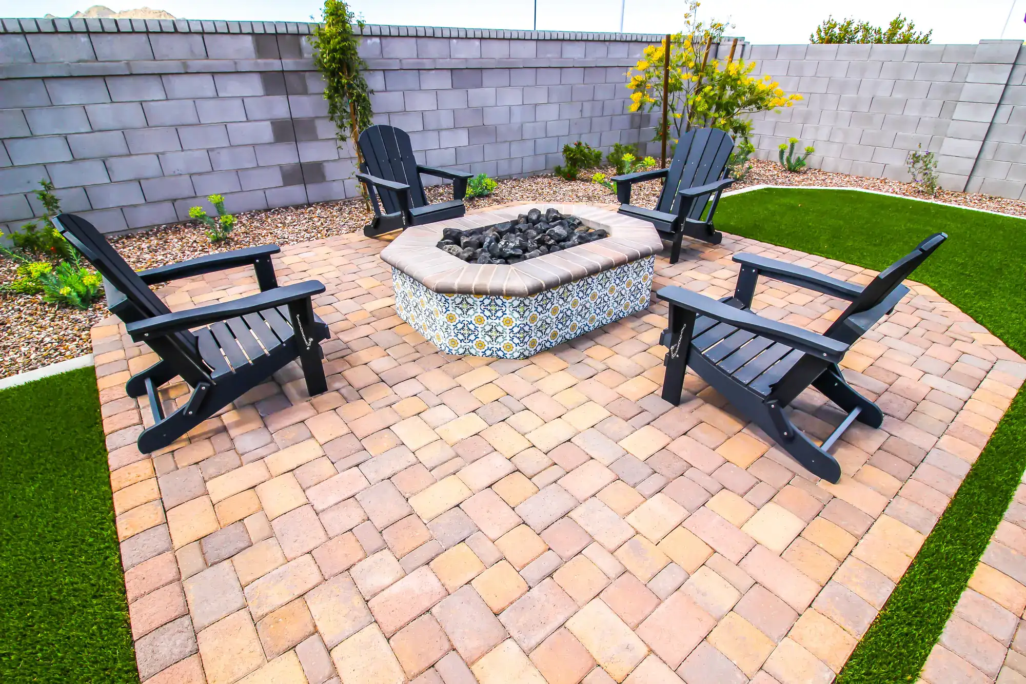 A backyard patio by a masonry contractor Long Island, NY, features four dark Adirondack chairs around a square fire pit, set on stone pavers with green artificial grass, gravel, and lush plants along a concrete block wall.