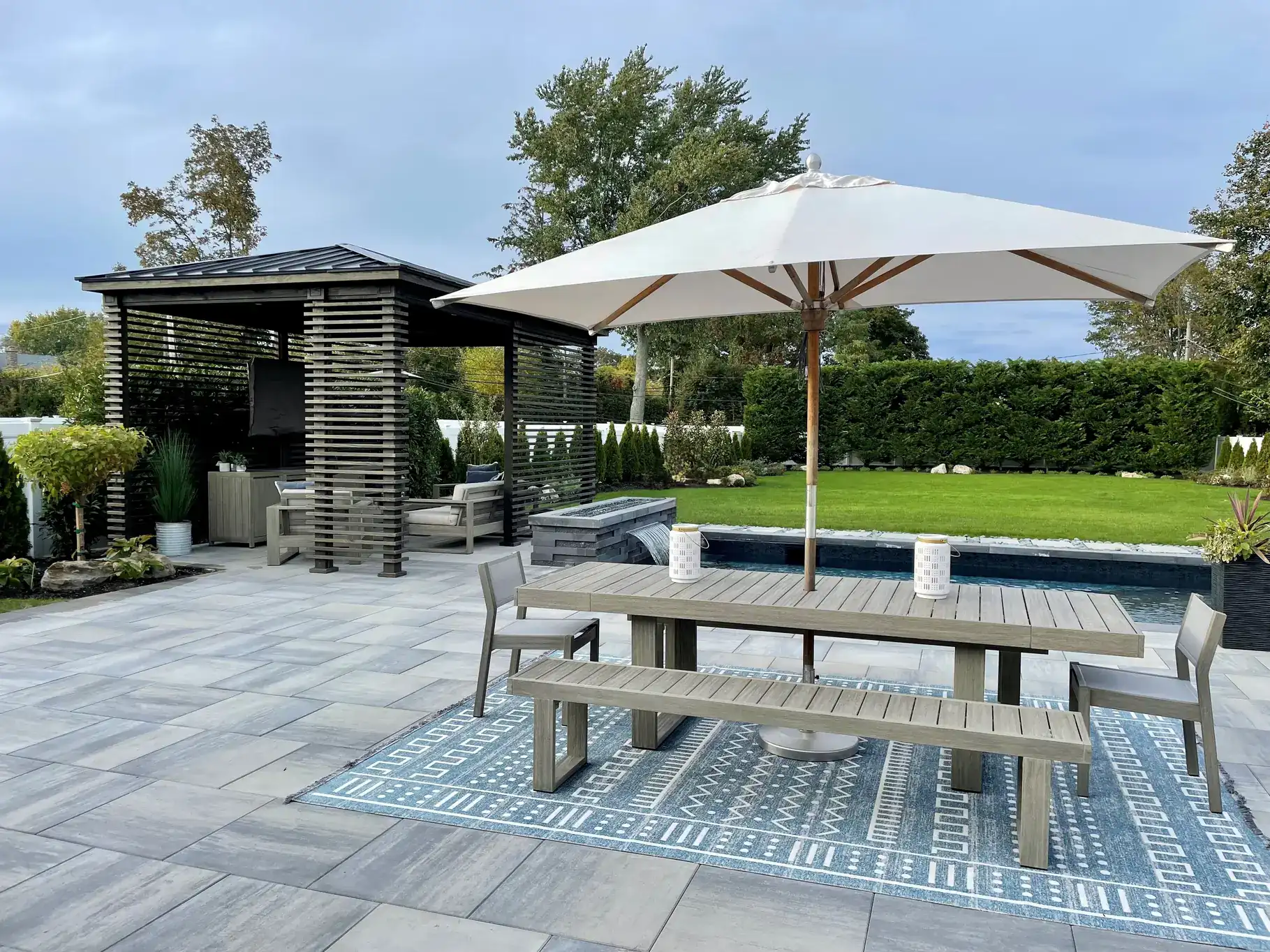 A modern outdoor patio by a top masonry contractor Long Island features a wooden picnic table, benches, and two candles under a large white umbrella on a patterned rug. In the background, there's a shaded pergola, green lawn, and trimmed hedges.