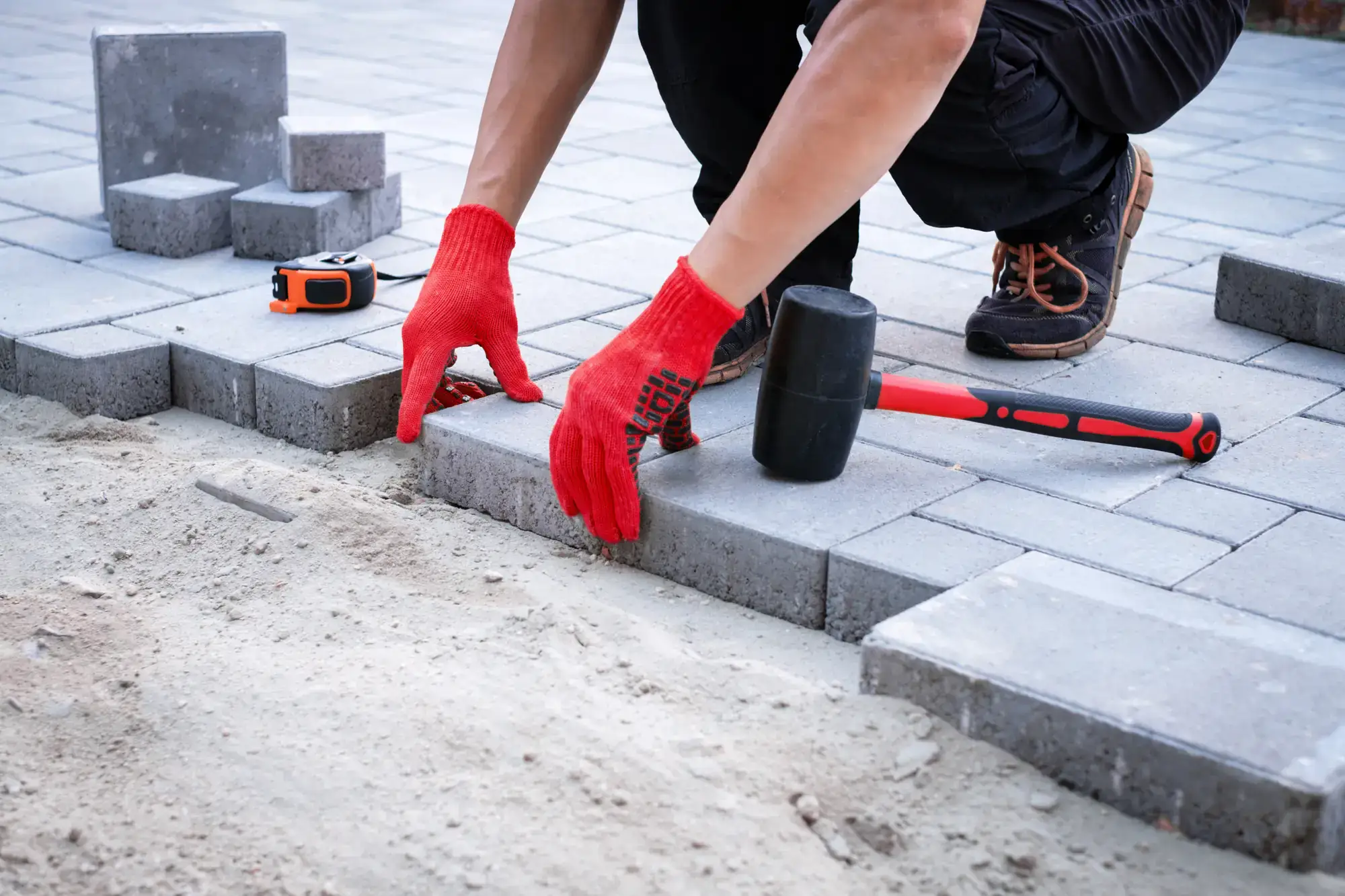 A masonry contractor Long Island in NY, wearing red gloves and black shoes, is laying gray paving stones with a rubber mallet and tape measure on a partially completed outdoor surface with sand in the foreground.
