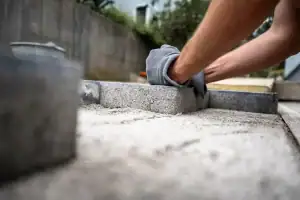 A masonry contractor in Long Island, NY, wearing gloves, positions a paving stone onto sand while laying a stone pathway or patio outdoors.