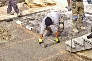 A worker in gloves and kneepads, part of a skilled masonry contractor Long Island, NY team, carefully places a large paving stone onto a pathway, while another person stands nearby on already laid pavers surrounded by construction materials.