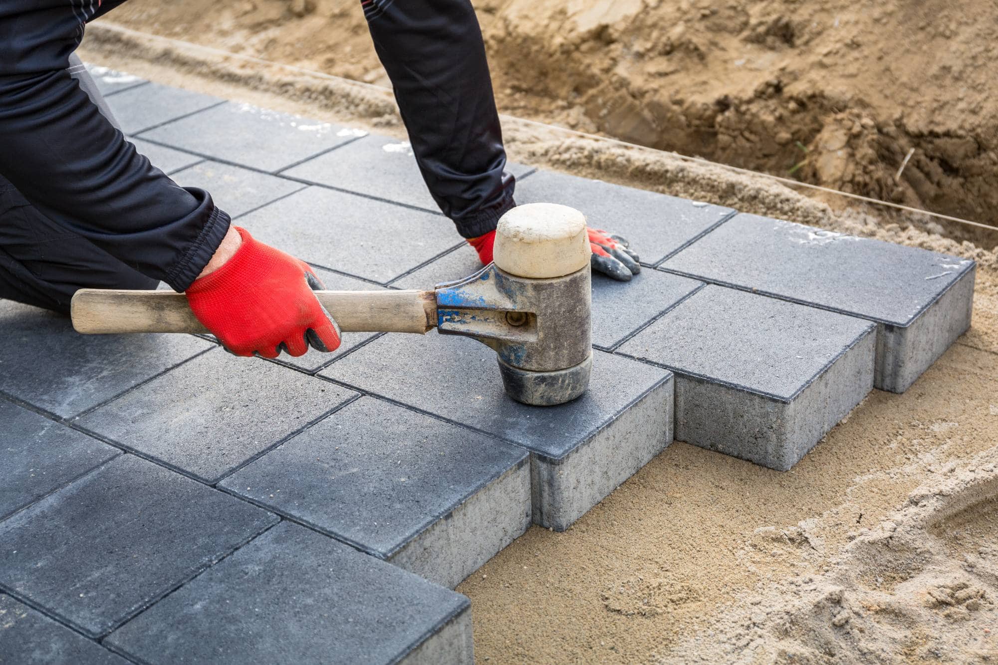 A masonry contractor in Long Island, NY, wearing red gloves, uses a rubber mallet to lay gray concrete paving stones on a bed of sand during walkway construction.