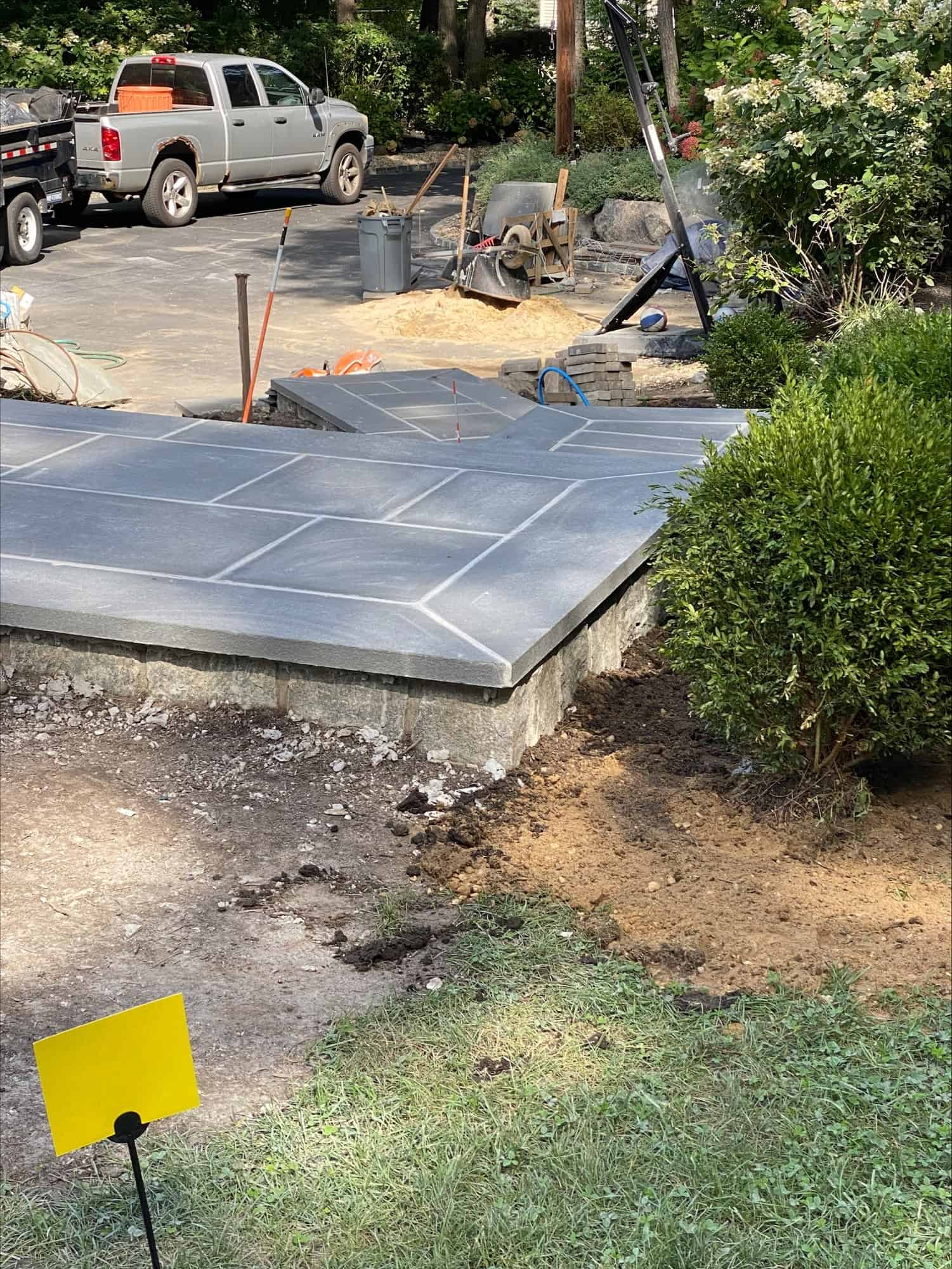 A newly constructed stone patio with large gray tiles is shown in a yard under renovation. Construction tools and materials are visible nearby, and a truck is parked in the background. Bushes and grass border the area.