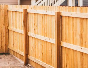 A close-up view of a light-colored wooden privacy fence with vertical planks and horizontal support beams in a backyard. The fence runs alongside a sandy area and a house with visible stairs in the background.