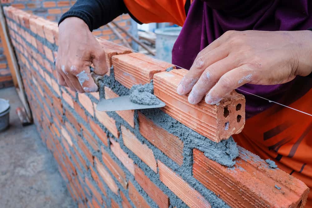 A person uses a trowel to spread mortar between red bricks while constructing a brick wall. The wall is partially built, and the worker's hands are visible placing a new brick in position.