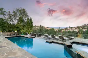 A luxurious outdoor pool with lounge chairs and a hot tub overlooks a scenic hillside neighborhood at sunset, surrounded by trees and stone decking.