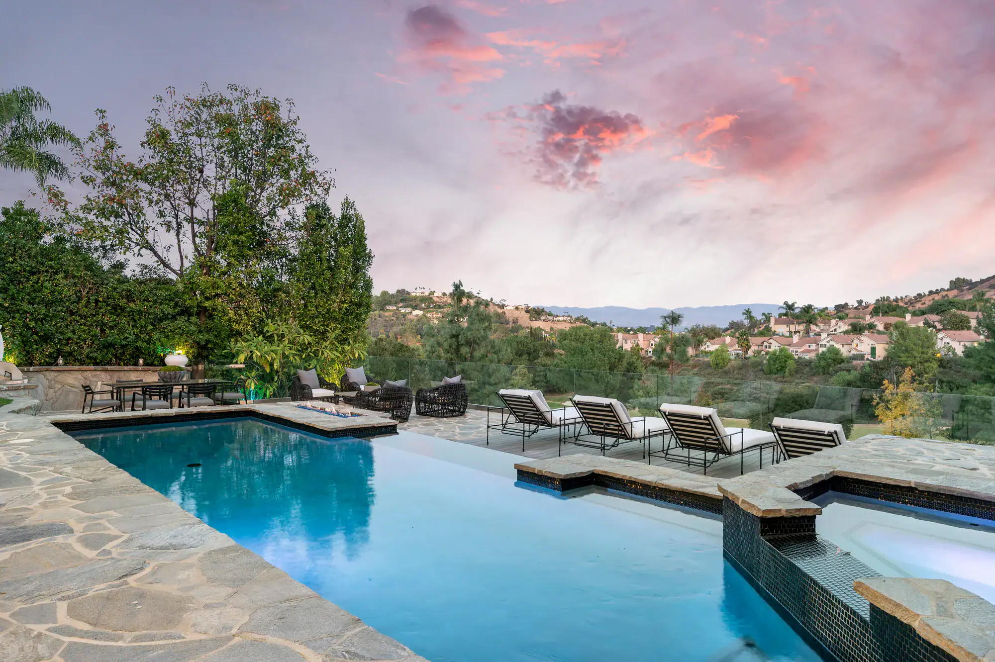 A luxurious outdoor pool with lounge chairs and a hot tub overlooks a scenic hillside neighborhood at sunset, surrounded by trees and stone decking.