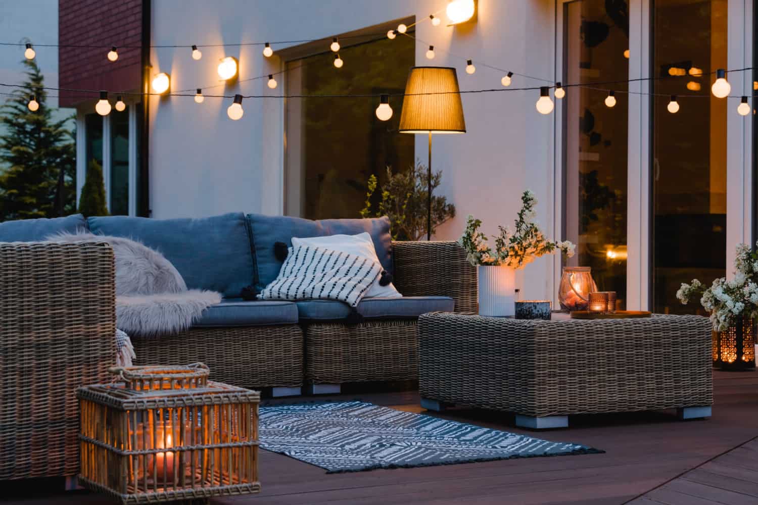 Outdoor patio with wicker furniture, a gray cushioned sofa, patterned pillows, a coffee table, lanterns, string lights, potted plants, and a floor lamp, creating a cozy, inviting atmosphere in the evening.
