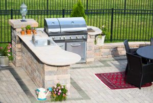 Outdoor patio with a built-in stone kitchen, featuring a grill, sink, potted plants, a dining table with chairs, a red rug, and a watering can, set on a paved surface with a black metal fence and grass in the background.