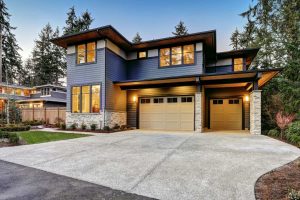 Modern two-story house with dark siding, large windows, stone accents, and a double garage, surrounded by trees and landscaped yard at dusk.