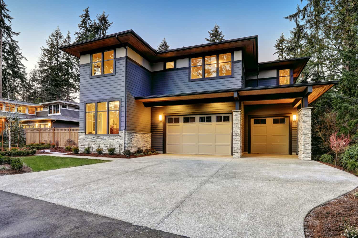 Modern two-story house with dark siding, large windows, stone accents, and a double garage, surrounded by trees and landscaped yard at dusk.