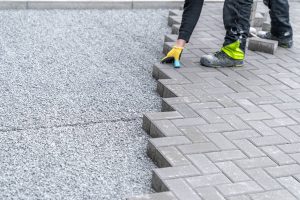A worker wearing gloves and protective boots is installing rectangular paving stones in a herringbone pattern on a gravel base for a pathway or patio.