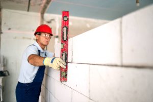 A construction worker wearing a red hard hat and gloves uses a red spirit level to check the alignment of a white brick wall under construction indoors.