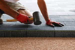 A person wearing red gloves and shorts uses a rubber mallet to align and secure gray paving stones over a bed of sand or gravel during outdoor construction.