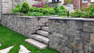 Stone steps lead up between two short retaining walls made of gray stone blocks, bordered by green grass and lush shrubbery, with a brick house partially visible in the background.