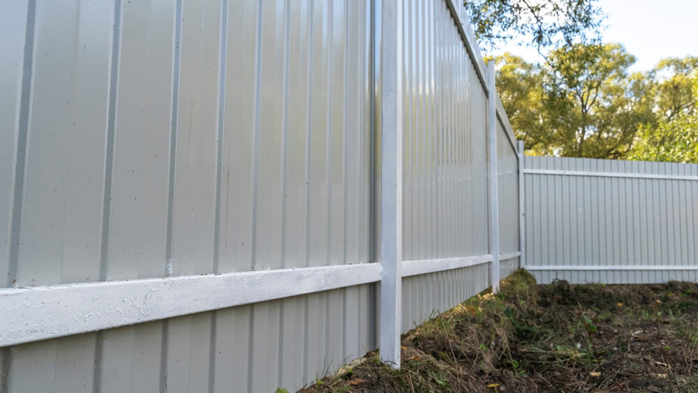 A close-up view of a gray metal fence with vertical panels and white support beams, set outdoors with trees and greenery visible in the background.