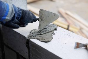 A person wearing a glove uses a trowel to spread wet cement on concrete blocks, likely while building a wall. Construction tools and materials are blurred in the background.