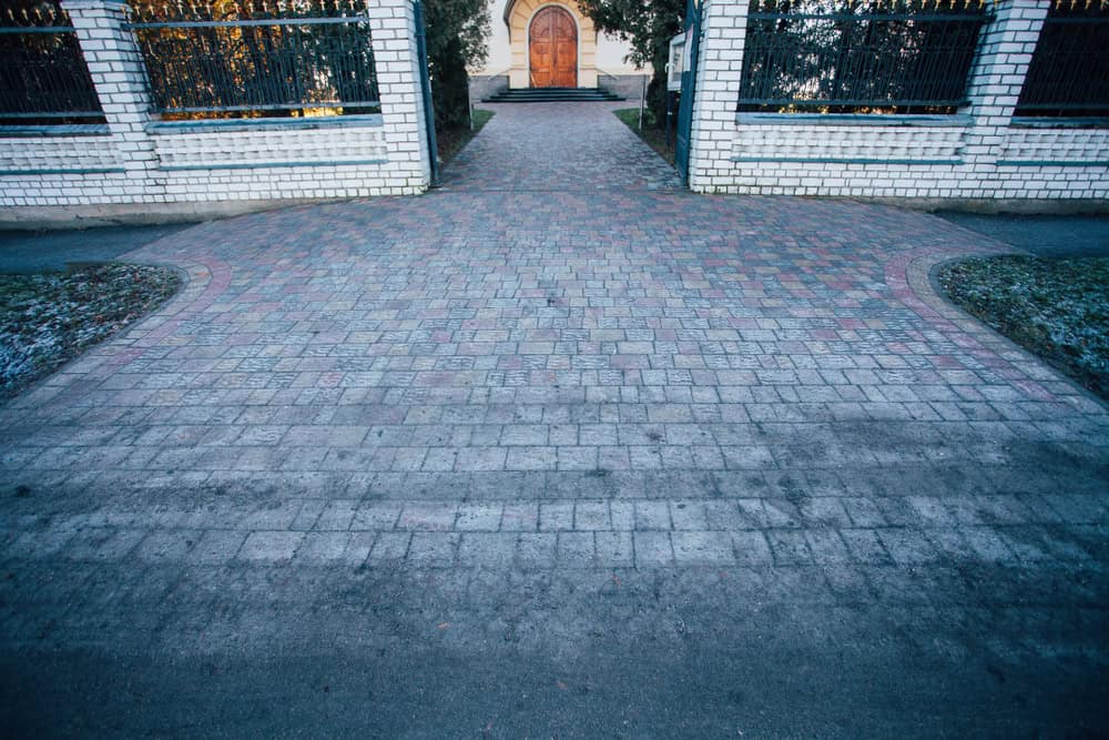 A paved walkway with a brick pattern leads to a wooden double door entrance in a white brick wall, flanked by iron fences and small patches of grass.