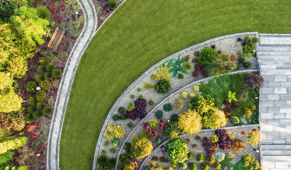 Aerial view of a landscaped garden with curved paths, green lawn, and tiered flower beds filled with various colorful plants, shrubs, and trees next to stone steps and a paved area.