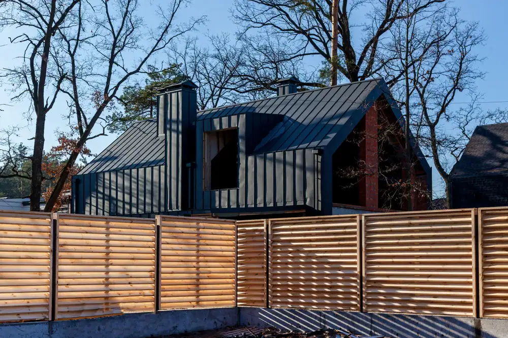 Modern house with a dark metal roof and exterior, visible behind a light wooden slat fence. Leafless trees surround the property, and the sky is clear and blue.