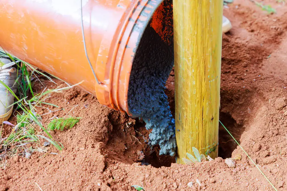 An orange bucket pours wet concrete into a hole in the ground around a wooden post, set in reddish soil with some green plants nearby.
